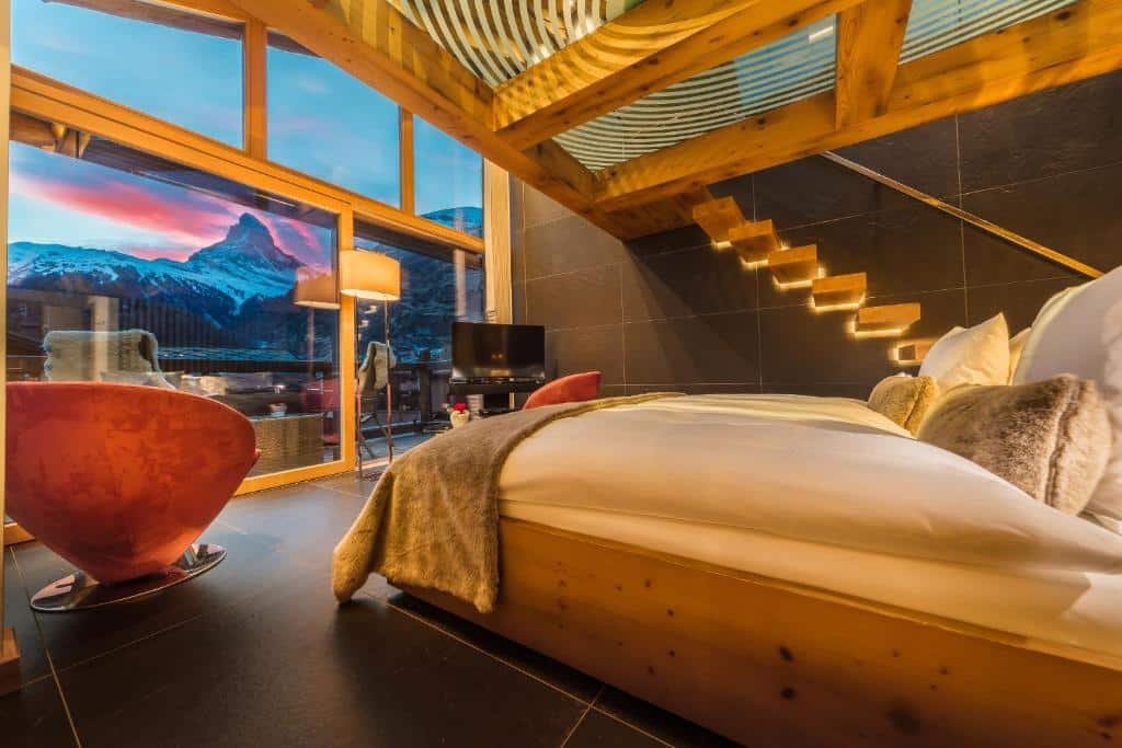 Quarto do Hotel Bellerive com uma cama de casal do lado direito da imagem do lado esquerdo da cama um poltrona e uma janela ampla com vista para as montanhas. 