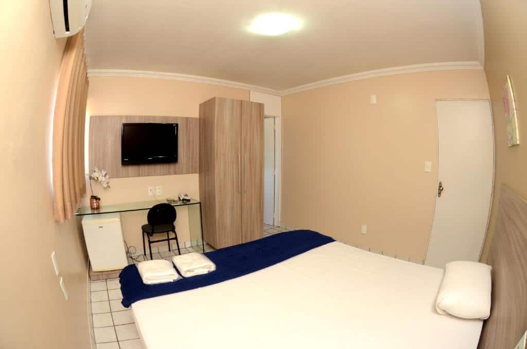 Quarto do Hotel Itaparica. A cama está centralizada, ao fundo há uma mesa de vidro, cadeira, frigobar, armário e uma TV presa a parede. No fundo do quarto e a direita há uma porta. Imagem para ilustrar o post hotéis em Caruaru.