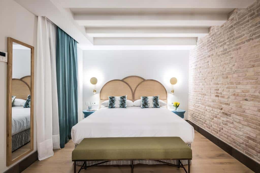 Quarto do Hotel Casa de Indias By Intur. Uma cama de casal com uma cabeceira e um banco na frente clean. Do lado esquerdo um espelho e a janela do quarto com cortina.