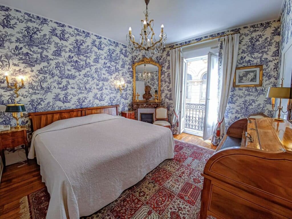 Quarto do Hôtel de l’Abeille com cama de casal do lado esquerdo da imagem com uma cômoda em cada lado da cama e a frente a cama um piano. Representa hotéis no Vale do Loire.