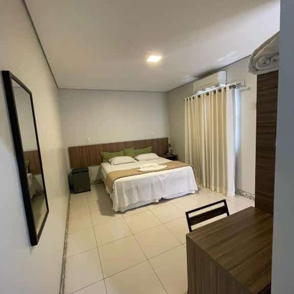 Quarto do Hotel e Pousada Oca com cama de casal no centro do quarto, com uma cômoda do lado direito perto da janela com cortinas brancas e frigobar do lado esquerdo, espelho  grande do lado esquerdo.