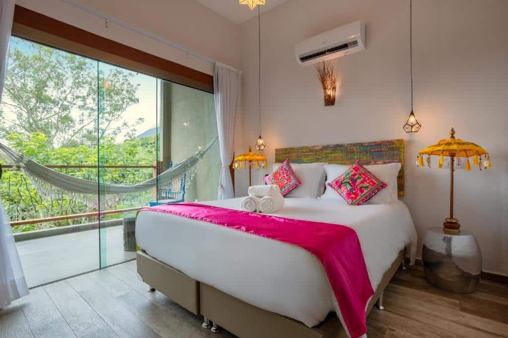 Foto do quarto em Hotel Vila Kebaya. Ilustra o post sobre Onde ficar em Ilhabela. Há uma cama box de casal no centro. Na parede atrás dela há um ar-condicionado e duas luminárias penduradas no teto. Dos dois lados da cama há um abajur. No lado esquerdo há uma porta de vidro com passagem para a varanda. Há uma rede na varanda.