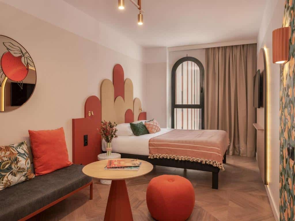 Quarto do Ibis Styles Sevilla City Santa Justa. Um sofá com uma mesa de centro e um puff na frente. Atrás a cama de casal com uma cabeceira colorida e de frente uma televisão. Atrás da cama a janela do quarto.