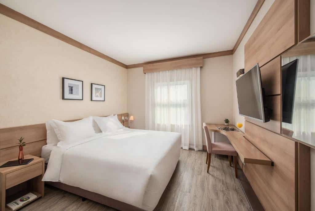 Quarto do Intercity Itupeva com chão de madeira, cama de casal, uma janela com cortinas, uma bancada de trabalho com uma cadeira e uma televisão