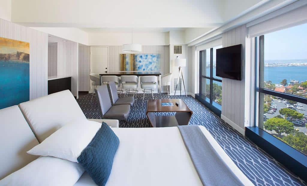 Quarto do Manchester Grand Hyatt San Diego com cama de casal com vista para a janela. Do lado esquerdo duas cadeiras com mesa de centro e TV a frente e ao fundo mesa com cinco lugares.