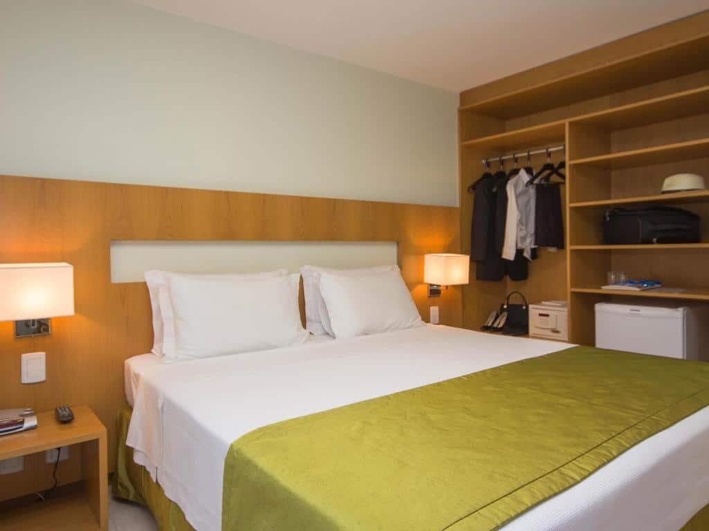 Quarto do Naoum Hotel com uma cama de casal, cabeceira com abajures presos, e um armário de conceito aberto do lado direito com lugar para pendurar roupas e outros itens pessoas, há também um frigobar dentro do móvel, para representar os hotéis perto do consulado americano em Brasília