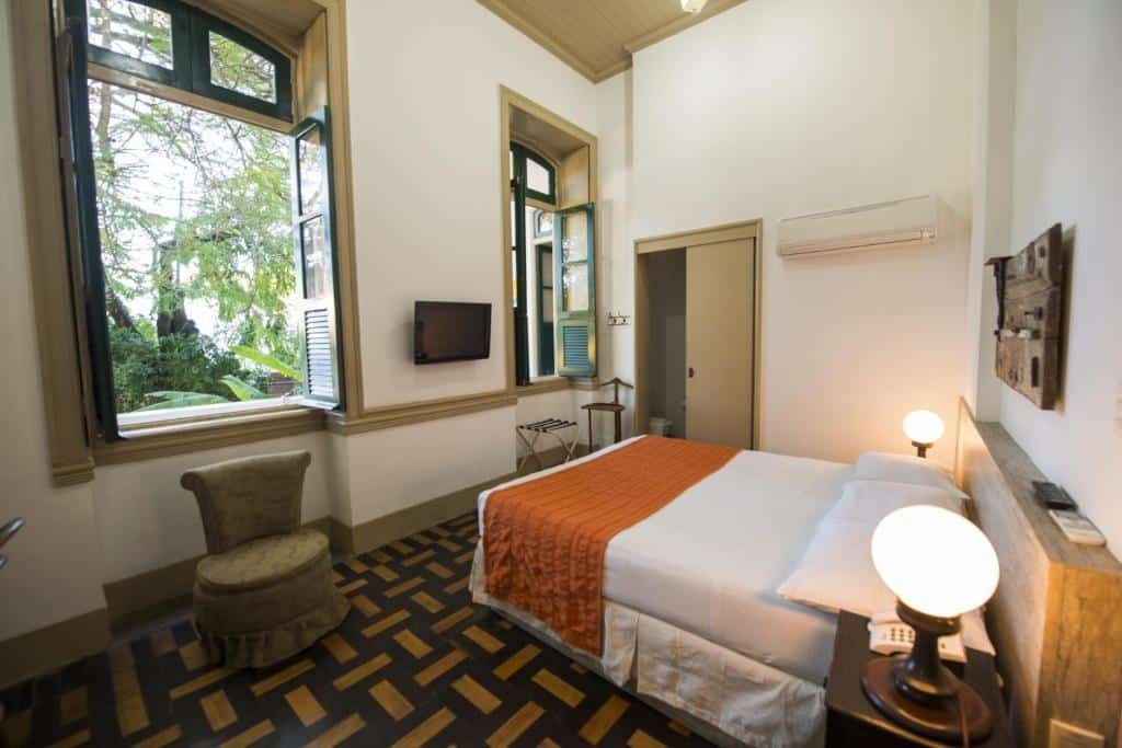 Uma cama com cômoda e abajur em cada lado. Na frente duas janelas. No quarto uma TV, uma poltrona e um ar-condicionado.