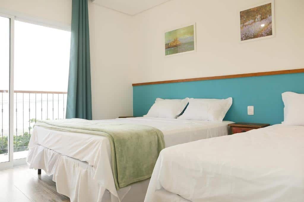 Quarto da Pousada Conca di Mare com uma cama de casal e ao lado uma cama de solteiro. De cada lado da cama de casal tem uma mesa de cabeceira e dois quadros na parede. Já do lado esquerdo da cama de casal tem uma porta de vidro com cortinas que dá acesso a varanda do quarto.