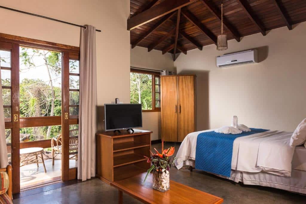 Quarto da Pousada Ilhote da Prainha. A direita é possível ver uma cama de casal, um armário de duas portas e uma janela com vista para a floresta. Na esquerda observa-se uma televisão em cima de rack de madeira, a frente da televisão uma pequena mesa baixa com planta em cima e uma porta com cortinas, que vai para a varanda. Imagem utilizada para ilustrar o post pousadas pet friendly em Ilhabela.