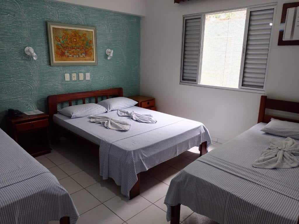 Quarto da Pousada Porto do Itagua com três camas, uma cama de casal e duas de solteiro. De cada lado da cama de casal tem uma mesa de cabeceira. No quarto tem também uma janela aberta.