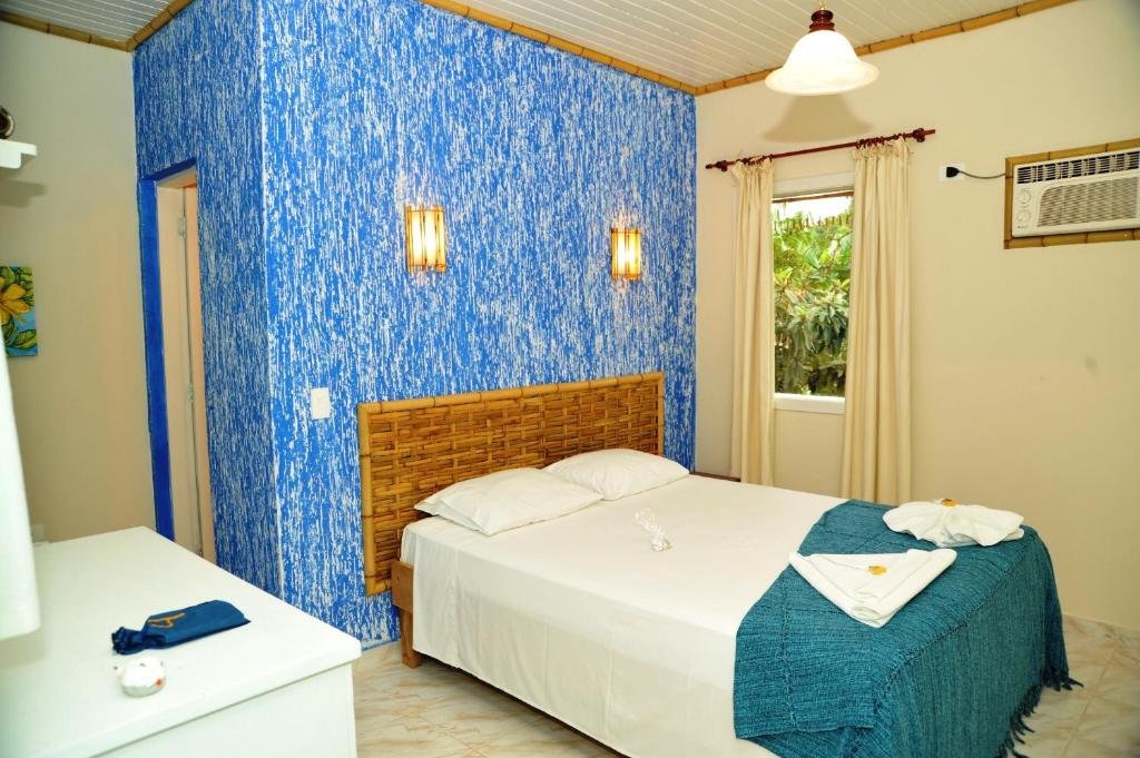 Quarto da Pousada Praia do Curral que mostra uma cama de casal e do lado esquerdo uma janela aberta com cortinas e um antigo ar-condicionado. Do outro lado da cama tem um móvel e uma porta que dá acesso a outra parte do quarto.