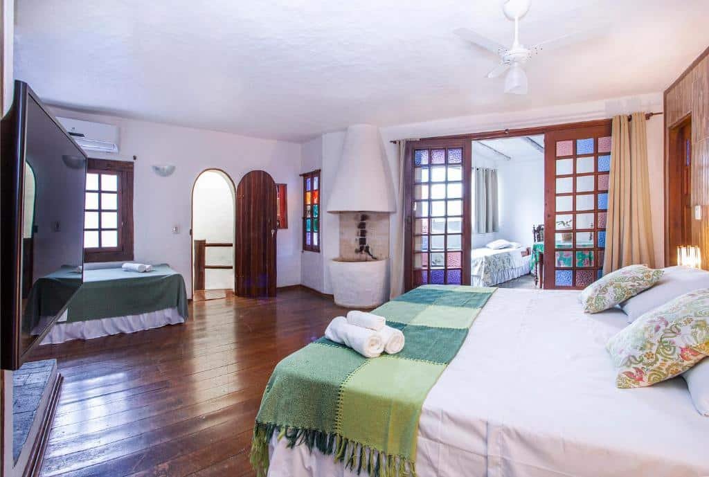 Quarto da Pousada Recanto da Villa. No lado direito há uma cama de casal. Ainda a direita, porém ao fundo há uma porta com cortina e vitrais, que vai para outro ambiente em que há uma mesa e cama de solteiro. No lado esquerdo está uma televisão e no fundo do lado esquerdo há uma cama de solteiro, uma janela e uma porta de saída. Imagem utilizada para ilustrar o post pousadas pet friendly em Ilhabela.