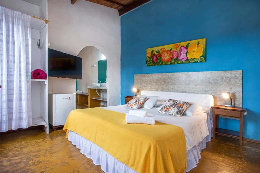 Quarto da Pousada Recanto da Villa que mostra uma cama de casal e de cada lado da cama tem uma mesa de cabeceira com um abajur. Já do lado esquerdo do quarto tem um guarda-roupa, um frigobar e uma tv e do lado um espaço aberto que dá acesso ao banheiro do quarto. 