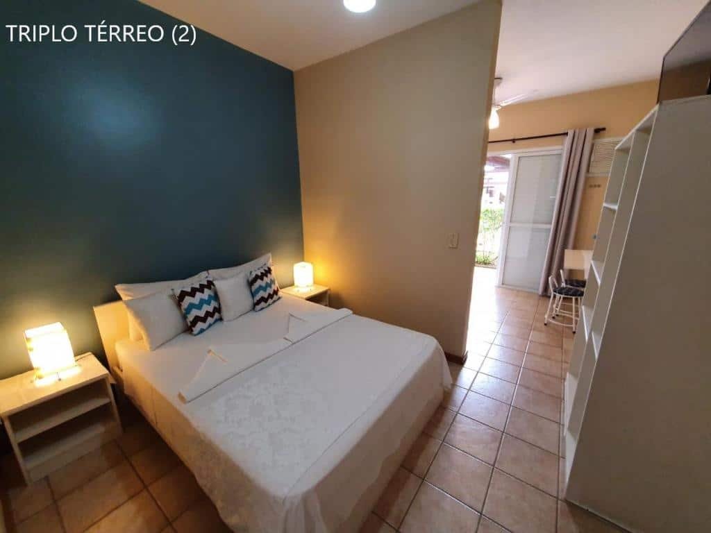 Quarto da Pousada Rosa dos Ventos Juquehy que mostra uma cama de casal e de cada lado da cama tem uma mesa de cabeceira. Em frente a cama tem um guarda-roupa e do lado dele tem parte de uma porta aberta aparecendo.