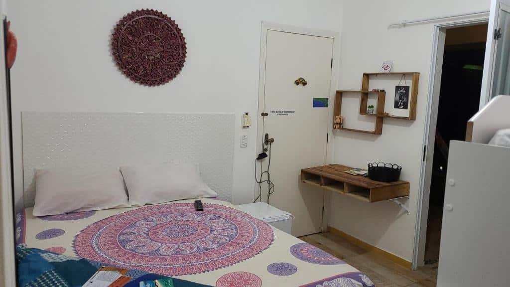 Quarto da Pousada Shiva com uma cama de casal e do lado direito um frigobar, uma porta, uma mesinha de madeira na parede e alguns nichos. Além disso, o quarto tem outra porta aberta que dá acesso a parte externa da pousada. 