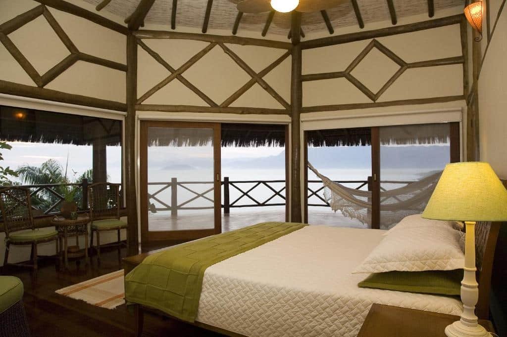Quarto da Pousada Singuitta. Na direita há uma cama de casal com abajur ao lado, na esquerda algumas cadeiras e uma pequena mesa. Ao fundo há portas que vão para a área externa do quarto, que têm rede, parapeito e vista para o mar. Imagem utilizada para ilustrar o post pousadas pet friendly em Ilhabela.