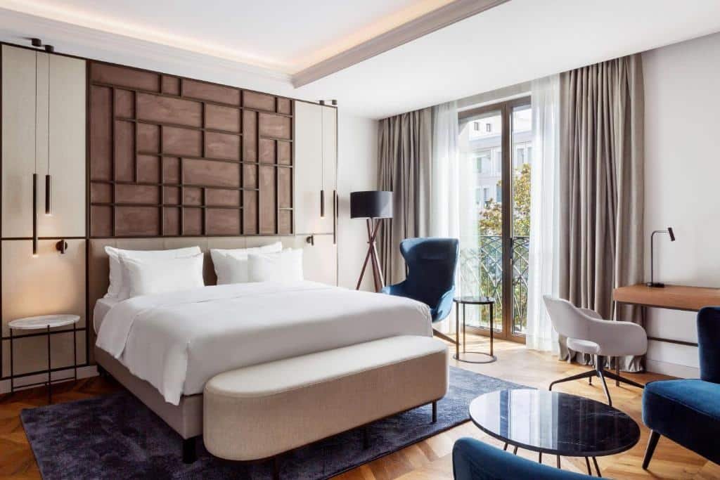 Quarto no Radisson Collection Hotel, Magdalena Plaza Sevilla. Uma cama de casal com um banco na frente no centro do quarto. De frente uma mesa com duas cadeiras. Do lado da cama uma poltrona e uma luminária, do lado da janela com vista pra rua. Do lado da janela uma mesa de trabalho. Foto para ilustrar post sobre Hotéis em Sevilha.