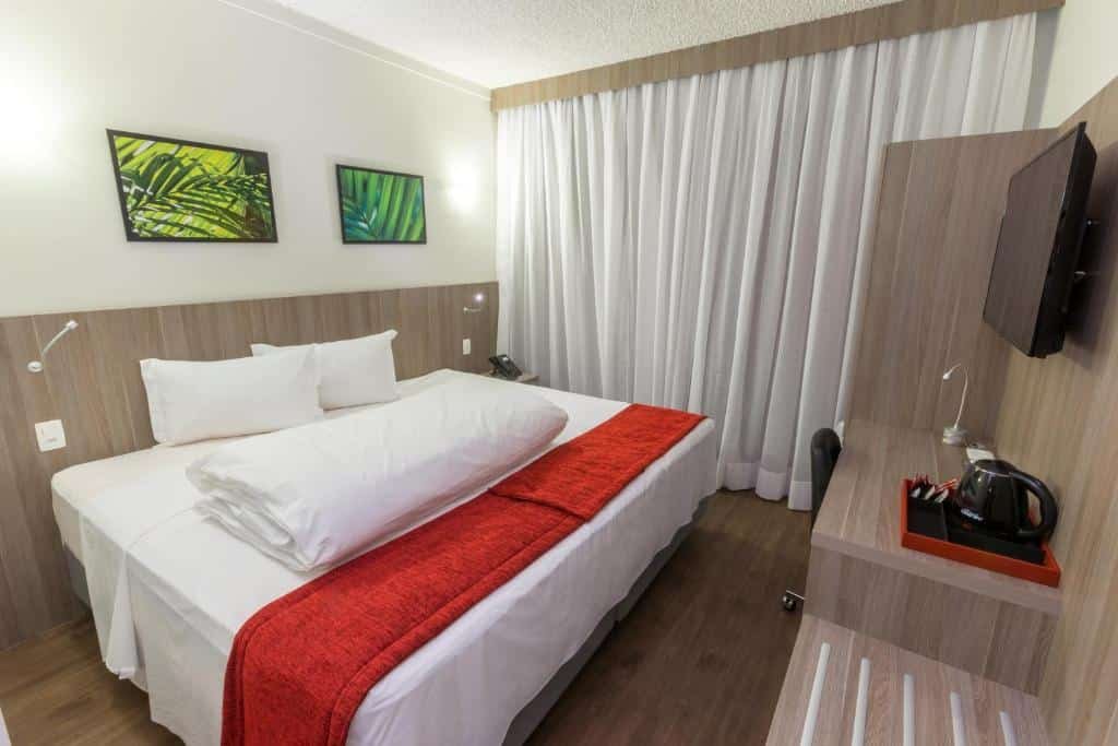 Quarto no Ramada by Wyndham Campinas Viracopos com uma cama de casal, uma janela com cortinas, uma mesa de escritório com uma cadeira, um armário de conceito aberto e uma televisão, para representar hotéis perto do Aeroporto de Viracopos em Campinas