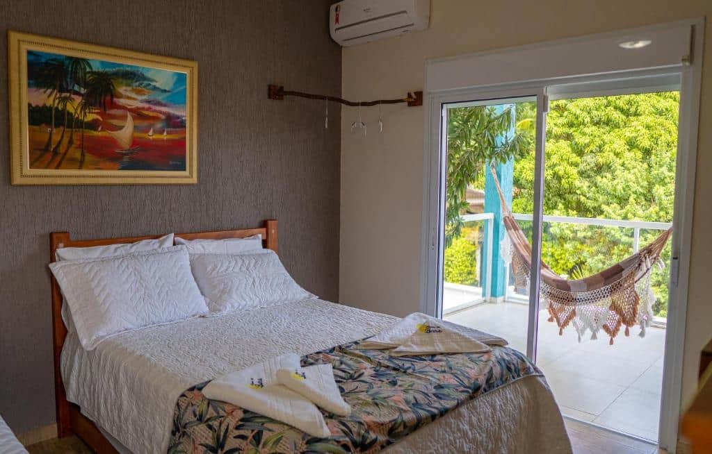 Quarto da pousada Residencial Águas de Bare com uma cama de casal, um quadro na parede, um cabideiro e um ar-condicionado. No quarto tem uma porta de vidro que dá acesso a varanda com uma rede e vista para as árvores.