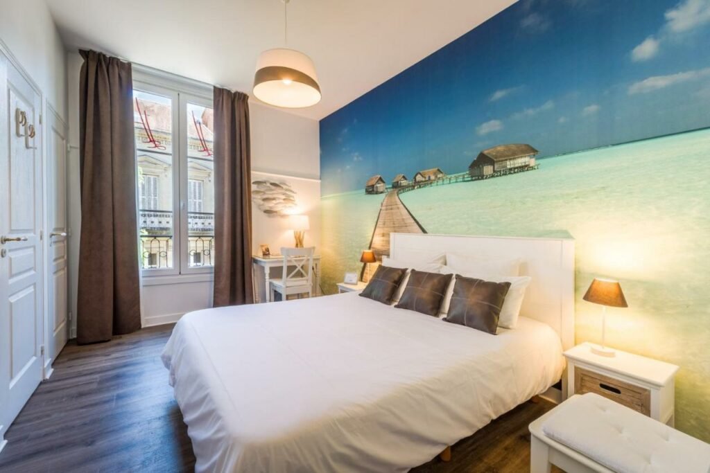 Quarto do The Originals Boutique, Hôtel Le Londres, Saumur (Qualys-Hotel) com cama de casal do lado direito da imagem com uma cômoda em cada lado da cama com luminária e do lado esquerdo da imagem uma mesa de trabalho com cadeira.