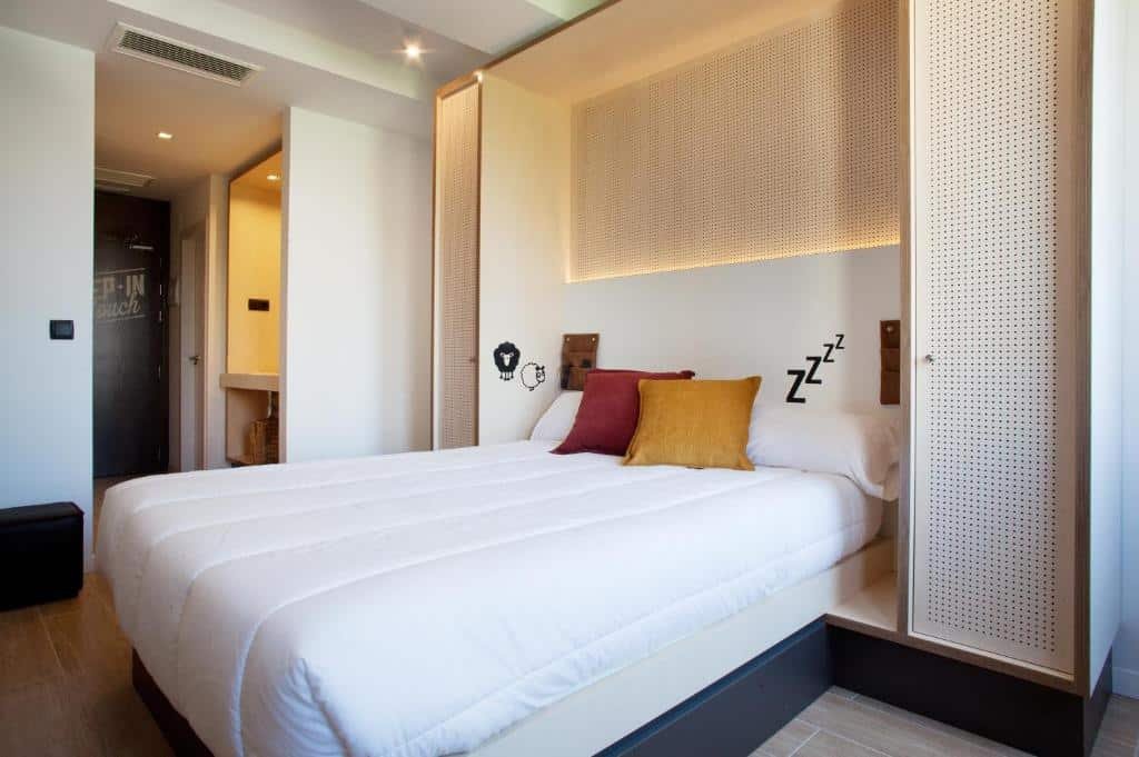 Quarto do Toc Hostel Sevilla. Uma cama de casal com travesseiros e duas almofadas em cima. Ao redor uma decoração clean, com um armário e uma porta de cada lado da cama. No fundo um corredor com a porta do quarto.