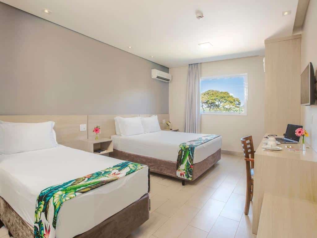Quarto do Valinhos Plaza Hotel com uma cama de casal, uma de solteiro, uma bancada de trabalho com uma cadeira, uma televisão e alguns itens para um café sob a bancada, para representar hotéis perto do Hopi Hari