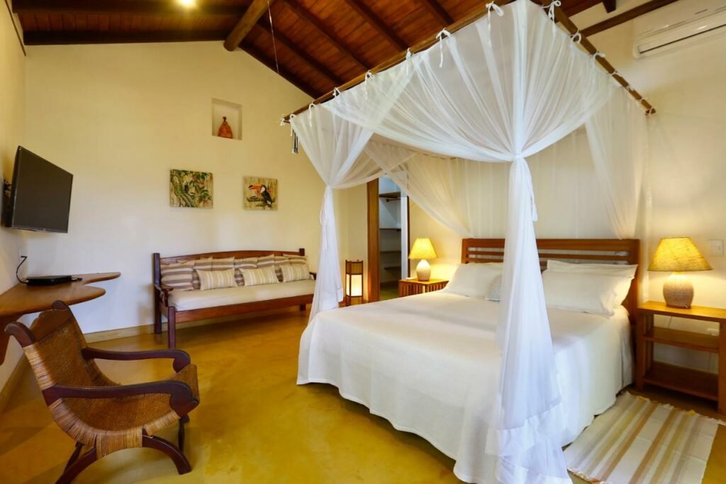 Quarto da Vila Monjuá Trancoso com cama de casal do lado direito da imagem com uma cômoda em cada lado com luminária, do lado esquerdo da cama um sofá e do lado esquerdo do sofá uma cômoda com TV presa na parede. Representa airbnb em Trancoso.