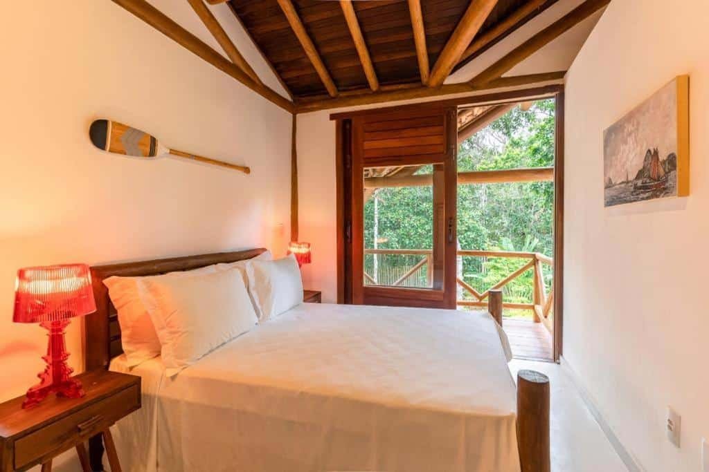 Quarto da Village Boutique La Dolce Vita com cama de casal do lado esquerdo da imagem e em cada lado da cama uma cômoda com luminária e do lado esquerdo uma porta com acesso a varanda.