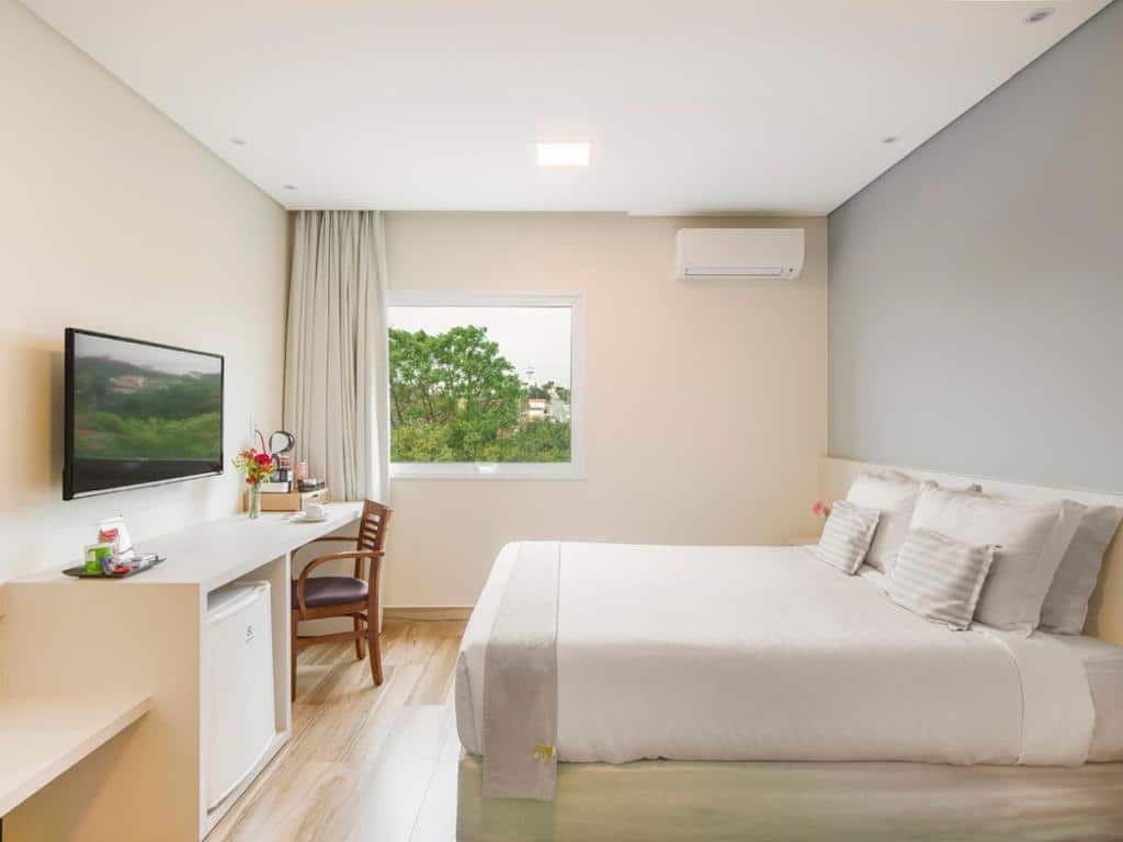 Quarto do Vinhedo Plaza Hotel com uma janela com cortinas, cama de casal, uma banca de trabalho com uma cafeteira e alguns itens de cozinha, uma cadeira, um frigobar e uma televisão, para representar hotéis perto do Hopi Hari