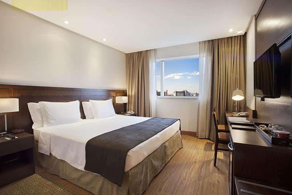 Quarto do Windsor Brasilia Hotel com chão de madeira, janela com cortinas, uma cama de casal e, de frente para ela, uma mesa de escritório com uma luminária e duas cadeiras, além de uma televisão, para representar hotéis perto do consulado americano em Brasília