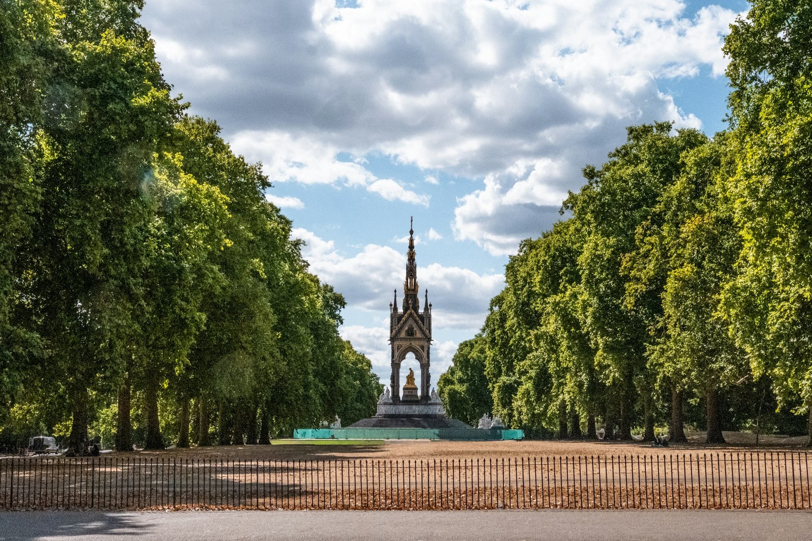 Um monumento em estilo gótico fica em um parque cercado por árvores, os Kensington Gardens, sob um céu parcialmente nublado, com uma cerca de ferro forjado em primeiro plano.