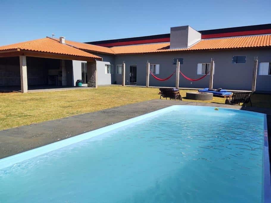 Parte externa do Recanto das Marias Casa com Fogueira com uma piscina grande do lado direto, do lado algumas cadeiras em volta da fogueira apagada e ao fundo a construção da casa com várias janelas e portas e uma área coberta com a churrasqueira.