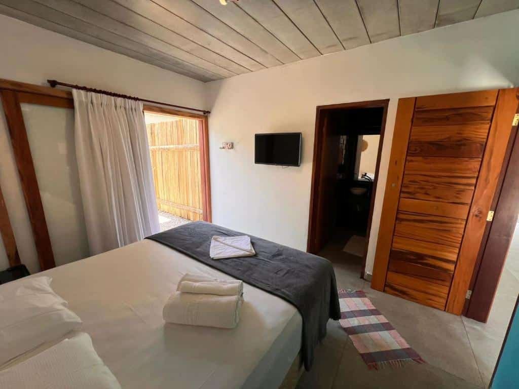 Reserva Ilhabela. Ilustra o post sobre hotéis em Ilhabela. Um quarto com uma cama, uma televisão na parede, cortinas e uma porta aberta que leva a outro cômodo. Aqui está uma descrição detalhada:

O quarto é iluminado e apresenta paredes brancas e um teto de madeira.
Há uma cama de casal com lençóis brancos e um cobertor cinza dobrado no pé da cama.
Toalhas brancas estão dobradas ordenadamente sobre o cobertor cinza.
Uma televisão preta está montada na parede à direita da cama.
Cortinas brancas cobrem uma janela de madeira à esquerda da imagem.
Um tapete colorido está parcialmente visível no chão em frente à cama.
Uma porta de madeira está aberta, revelando outro espaço não identificado do lado de fora do quarto.