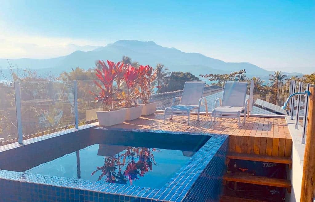Reserva Ilhabela. A foto mostra um cenário sereno e relaxante de um terraço ao ar livre com uma piscina. Aqui estão os detalhes:

A imagem apresenta um terraço com uma piscina.
A piscina tem bordas de azulejos azuis e reflete as plantas e o céu claro.
Duas cadeiras de descanso brancas estão posicionadas ao lado da piscina, convidando para o relaxamento.
Plantas vermelhas e verdes estão plantadas em vasos brancos modernos, adicionando um toque de cor vibrante à cena.
O terraço é cercado por um guarda-corpo de vidro transparente, proporcionando uma visão desobstruída das montanhas majestosas e do céu azul ao fundo.
O sol brilha intensamente, banhando o espaço em luz natural e destacando a beleza tranquila do ambiente.