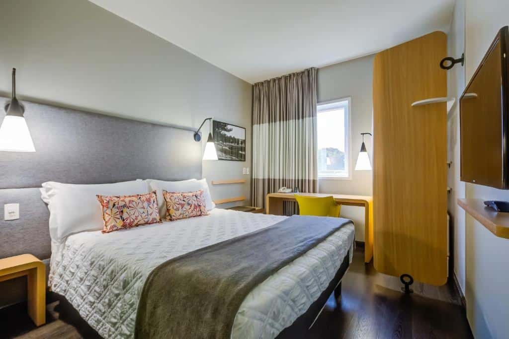 Quarto no Rio hotel by Bourbon Indaiatuba Viracopos com uma cama de casal, luminárias presas na cabeceira, uma janela com cortinas e uma mesa de escritório com uma cadeira, um armário com prateleiras e uma televisão