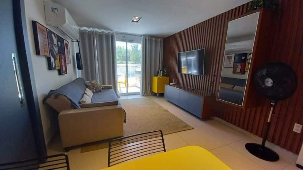 Sala de estar do Riviera Beach Place - Aquiraz Riviera. Um sofá está em cima de um tapete e de frente para uma tv e um espelho na parede. Ao fundo está a porta para a varanda. Em primeiro plano, no canto da imagem, é possível ver uma mesa com cadeiras.