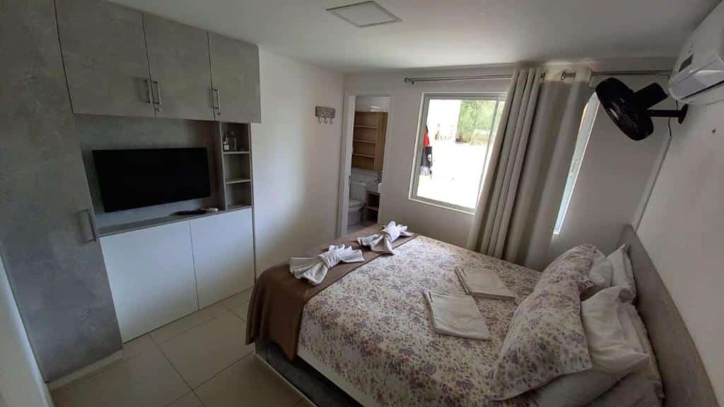 Quarto do Riviera Beach Place - Aquiraz Riviera. Uma cama de casal está no meio, de frente para uma tv e um armário. Na parede ao lado está um banheiro e uma janela, além de um ventilador e um ar-condiconado perto do teto.