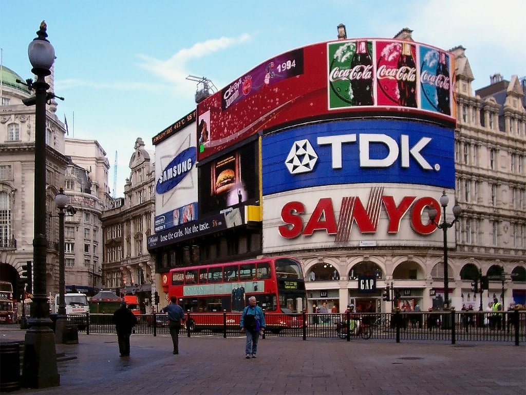Piccadilly Circus em Londres, com grandes anúncios digitais e um ônibus vermelho de dois andares em meio a prédios.