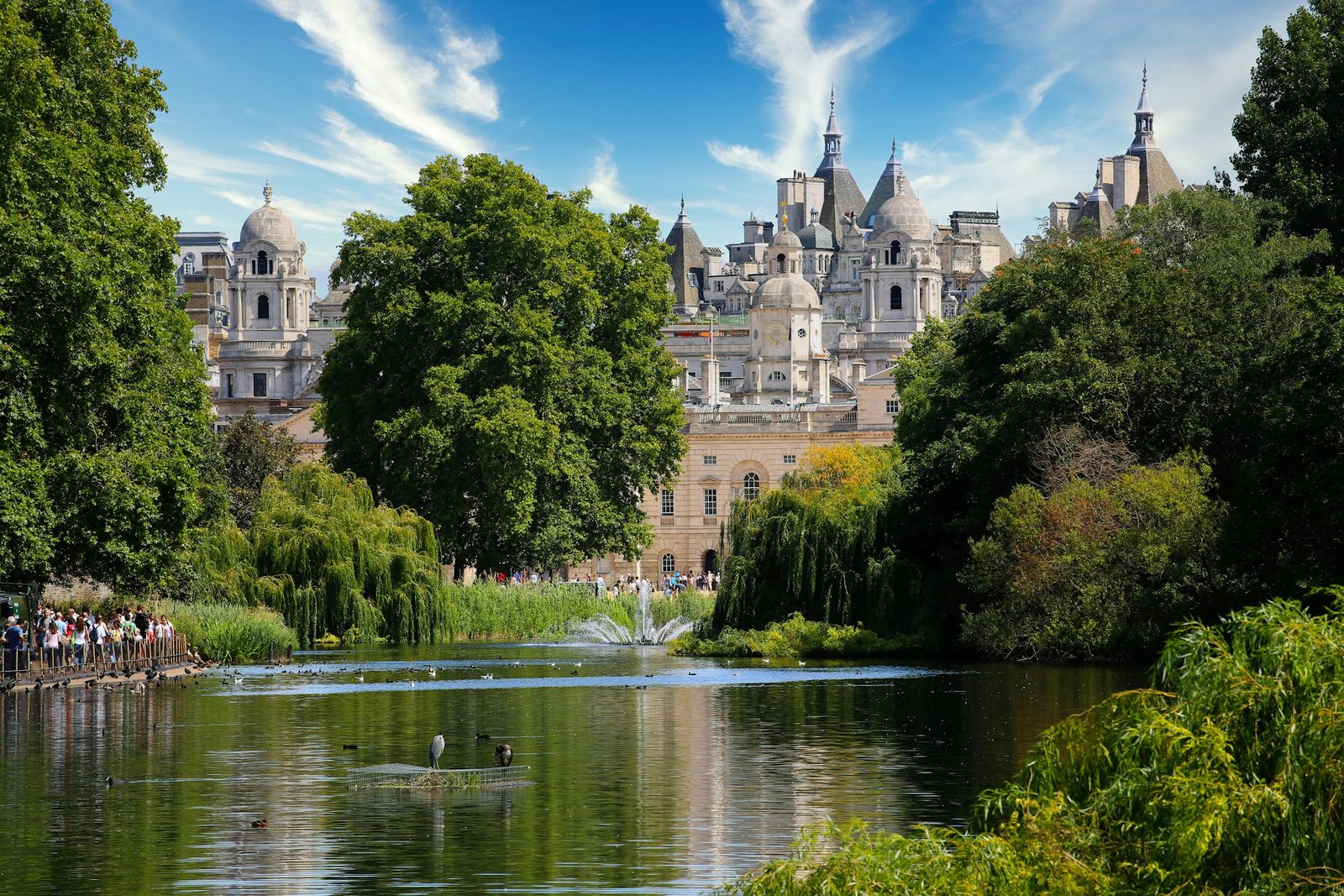 Um parque com um lago, cercado por árvores e edifícios com cúpulas e torres ao fundo, sob um céu azul com nuvens.