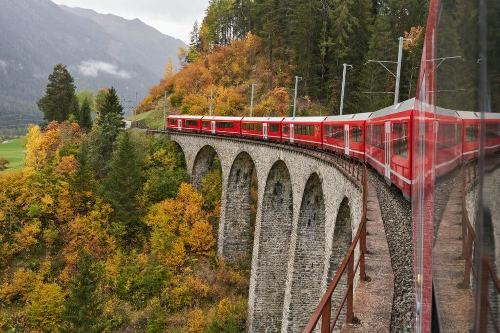 Um trem vermelho em Albula viaja por um viaduto de pedra curvo cercado por folhagens de outono com montanhas ao fundo. Representa como viajar de trem pela Suíça.