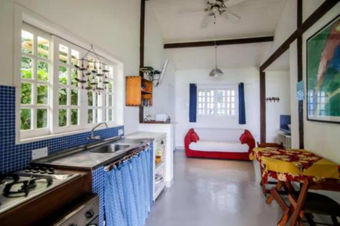 Cozinha das Suites e Bungalow Sol-Lua. Encostados na parede esquerda estão a pia, o fogão, o frigobar e os armários com utensílios. Uma janela fica logo acima da pia. Na parede oposta há uma mesinha com duas cadeiras. Ao fundo há um sofá abaixo de outra janela.
