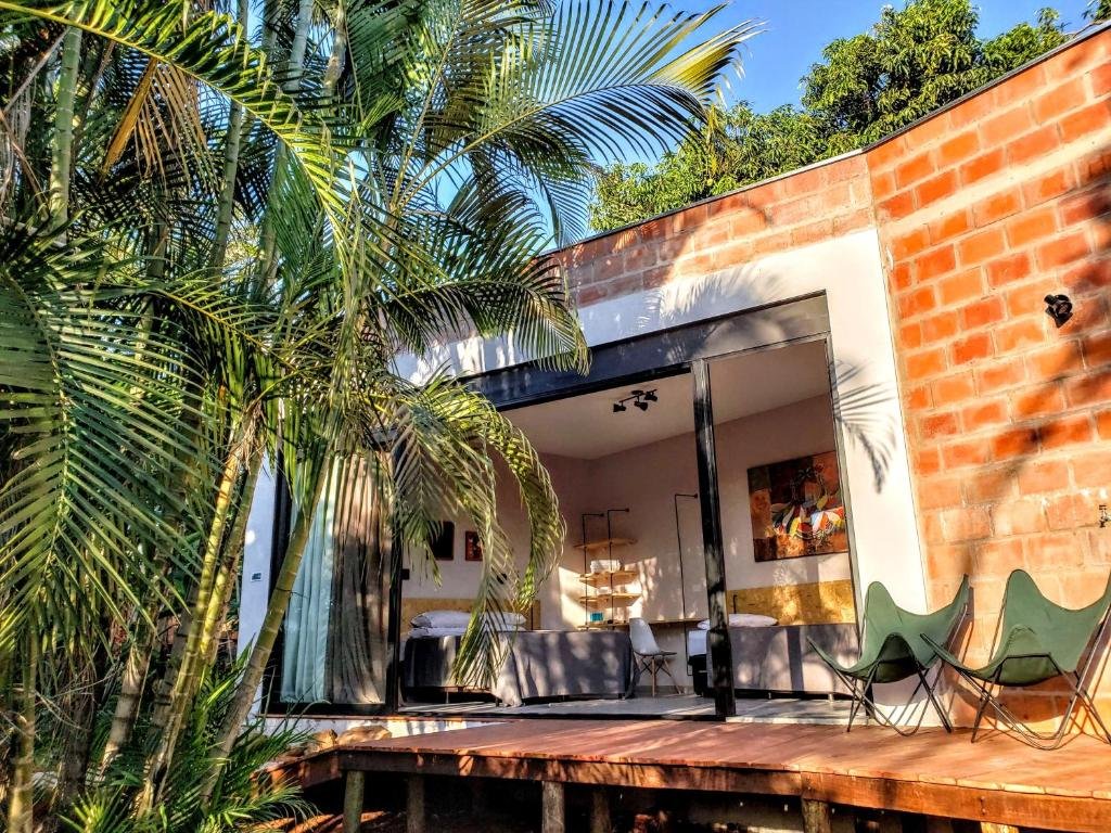 Fachada de tijolos do quarto do Suítes no Quintal que mostra uma palmeira e uma rampa de madeira que dá acesso a uma pequena varanda de madeira com duas cadeiras e uma porta grande vidro que separa a varanda do quarto. Imagem ilustrando post Airbnb em Brotas.