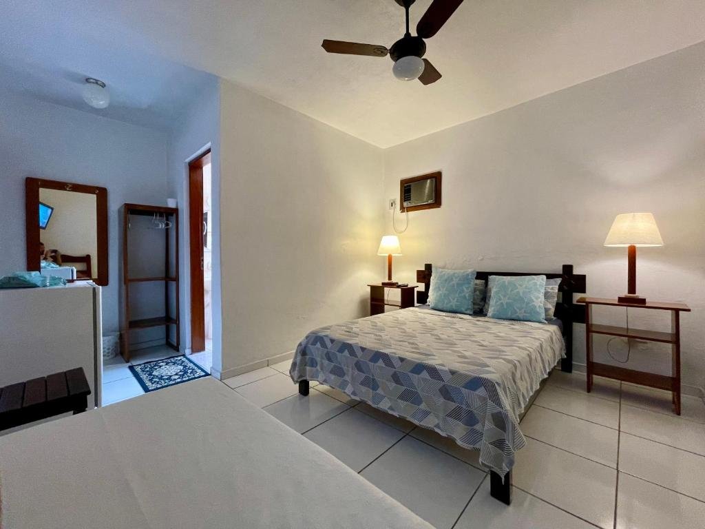 Suítes Nova Mar Ilhabela. Ilustra o post sobre hotéis em Ilhabela. Paredes brancas e piso de azulejos claros.
Uma cama de casal com uma colcha estampada em tons de cinza e azul, acompanhada por travesseiros combinando.
Mesas de cabeceira escuras de cada lado da cama, com abajures acesos em cima.
Um ventilador de teto instalado no centro do teto branco.
À esquerda da imagem, uma porta aberta revela outra sala ou corredor adjacente ao quarto principal. É possível ver outro móvel e um tapete azul escuro através da porta aberta.