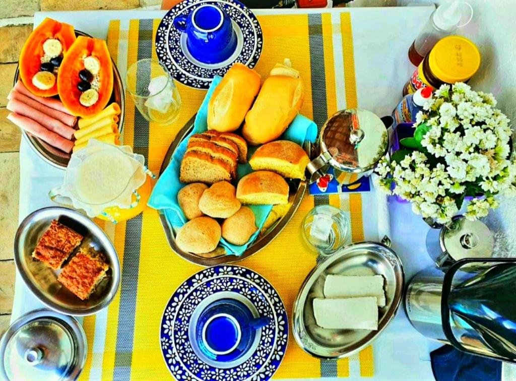 Suítes Nova Mar Ilhabela. A imagem apresenta uma mesa colorida posta para o café da manhã.
Há um tecido listrado amarelo e branco cobrindo a mesa.
Uma variedade de pratos e tigelas contendo alimentos estão dispostos sobre a mesa:
Um prato com fatias de mamão, bananas e outras frutas.
Um prato com pães variados, incluindo pães redondos e baguetes.
Uma tigela azul decorada contendo uma bebida ou sopa.
Um recipiente com algo que parece ser queijo ou tofu.
Outro recipiente metálico contendo pedaços retangulares grelhados de um item alimentar não identificado.
Há também embalagens de condimentos visíveis ao lado dos pratos, mas os rótulos não são claramente visíveis para identificar o conteúdo específico.
Uma jarra transparente possivelmente contendo água ou outra bebida clara está presente na mesa.
Flores brancas em um vaso adicionam um toque decorativo à configuração.