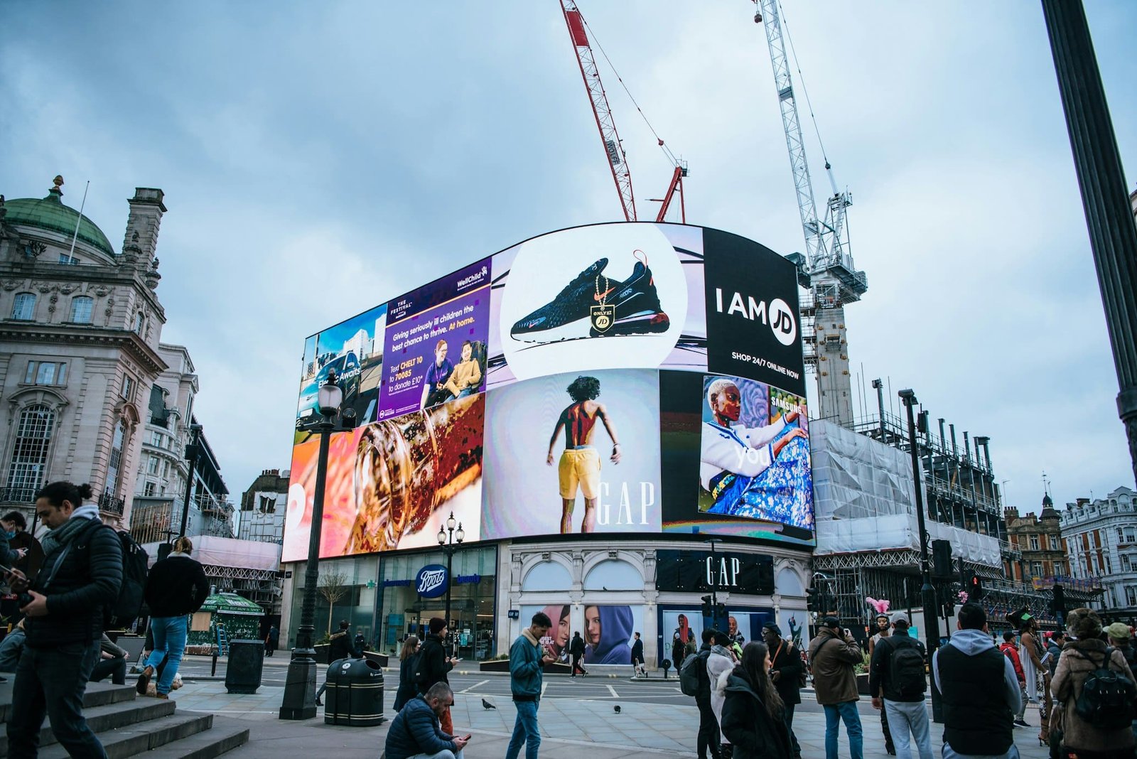 Pessoas caminham por uma movimentada praça da cidade com grandes anúncios digitais em uma tela curva e guindastes ao fundo. Esse é o Piccadilly Circus, um dos pontos turísticos de Londres.
