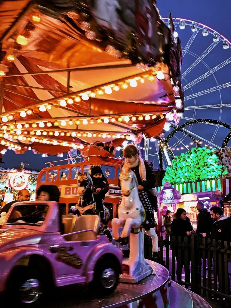 Crianças andam no carrossel do Hyde Park com um cavalo e um carrinho roxo, cercados por luzes festivas e uma roda gigante ao fundo.