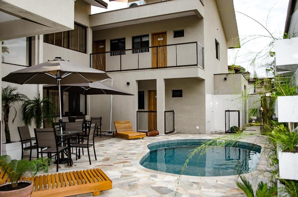 Parte da pousada Tons de Mar Residence que mostra do lado direito uma piscina e uma parede com vasos de plantas verdes. Já do lado esquerdo tem duas espreguiçadeiras de madeira, duas mesas com guarda-sol e cadeiras, e em volta a construção de dois andares da pousada. Imagem ilustrando post Pousadas pet friendly no Litoral Norte de São Paulo.