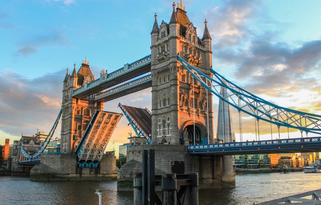 Tower Bridge, um dos pontos turísticos de Londres, com suas basculantes levantadas, sob um céu parcialmente nublado ao pôr do sol. O Rio Tâmisa é visível abaixo.