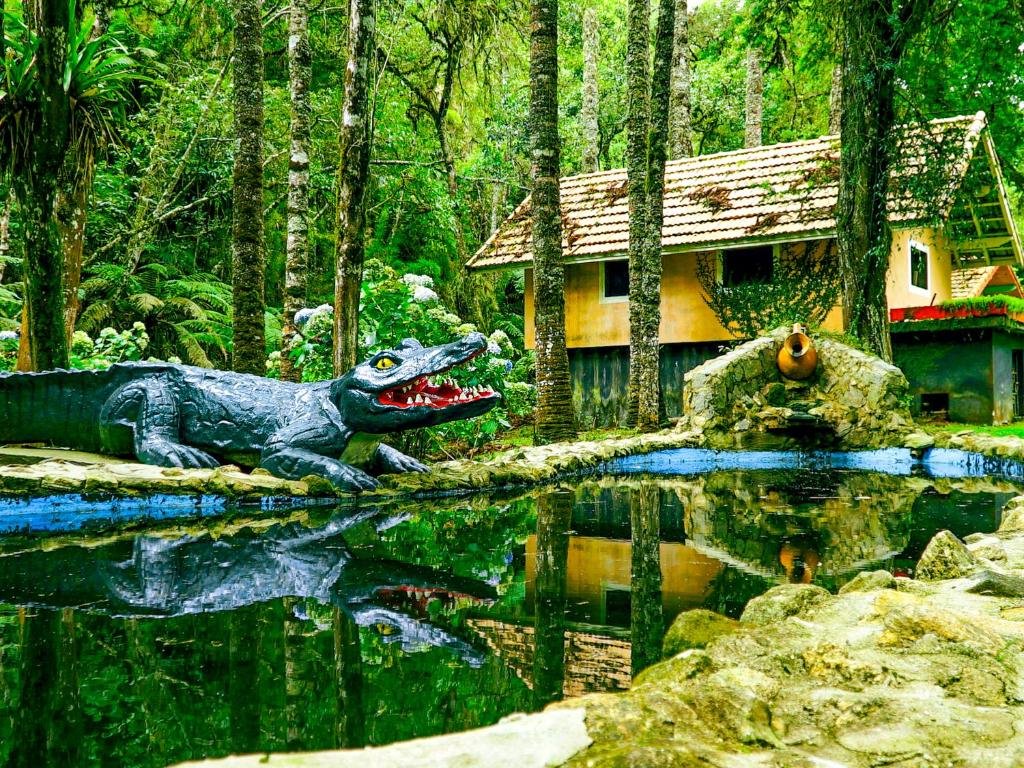 Foto da VELINN Áustria Hotel Monte Verde. Nessa foto, há uma escultura de um crocodilo com a boca aberta, posicionada em um lago cercado por pedras. Ao fundo, árvores altas e densas e uma casa com telhado de duas águas e paredes amarelas podem ser vistas. A cena evoca uma mistura de beleza natural e arte humana.