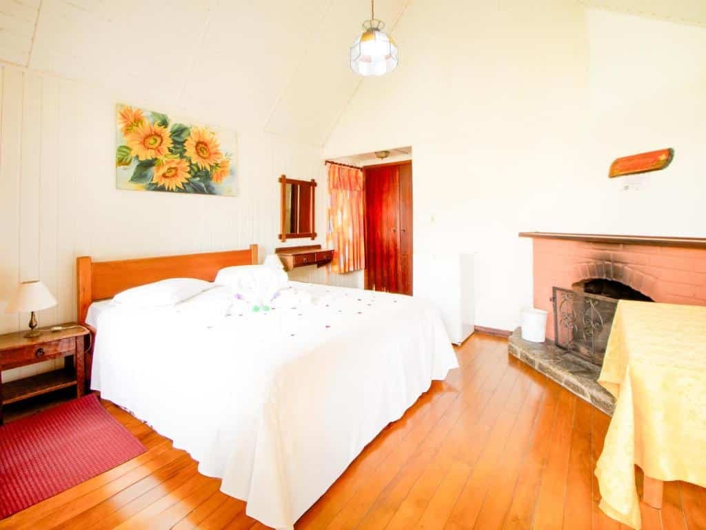 Foto da VELINN Áustria Hotel Monte Verde que está ilustrando o post de pousadas pet friendly em Minas Gerais. Nessa foto, há uma cama com lençóis brancos no centro, uma mesa de cabeceira à esquerda com um abajur, um quadro de girassóis na parede acima da cama, uma porta aberta à direita que leva a outro cômodo, e uma lareira com um espelho acima dela no canto direito da imagem.